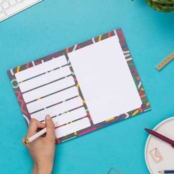 A4 Planner pad with black background, and multicoloured abstract shapes across it. This pad has 7 rectangular boxes in rows on the left of the pad, with one large white rectangular box on the right half. It is on a teal desk with a hand holding a pen to write on it, to the left.
