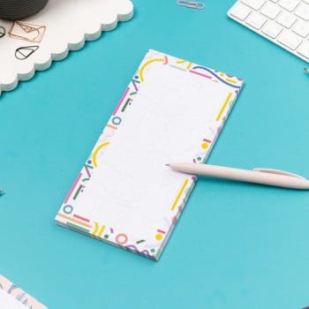 Cutouts List pad is on a teal desk, with a white pen leaning on it, with a white keyboard and paperclips around it.