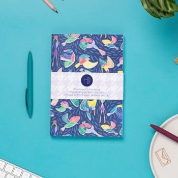 Paradise A5 Dot Grid Notebook, with its belly-band packaging around it, is on a teal desk with some pens, and a keyboard to the bottom left corner. The book's cover features a navy background with line-drawn bird motifs and offset coloured blocks behind them.