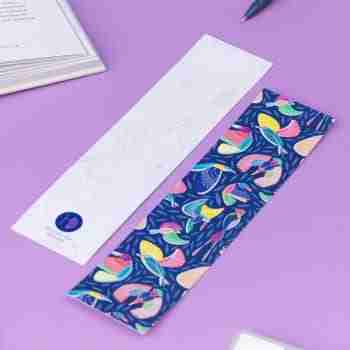 Two of the paradise double-sided bookmarks, on a lilac desk with a book open to the top left corner. One side features the full-colour patterned side of the Paradise pattern with the navy background, bird line motifs and colour blocks behind. The other side is the light-grey line drawing.