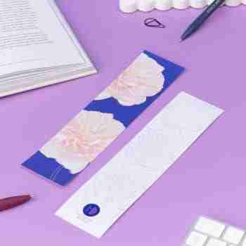 Two bookmarks, showing each side of the Spring Forward Bookmark. One side has a blue background with pink dhalia illustrations and golden ellow outlines, the other side is white with light grey outlines of the dahlia. They are on a lilac desk with an open book, pens and keyboard around them.